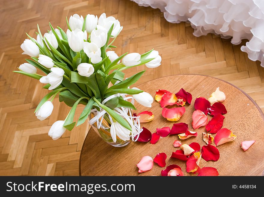 Vase with white tulips on a wooden little table and petals of a red tulip beside. Vase with white tulips on a wooden little table and petals of a red tulip beside