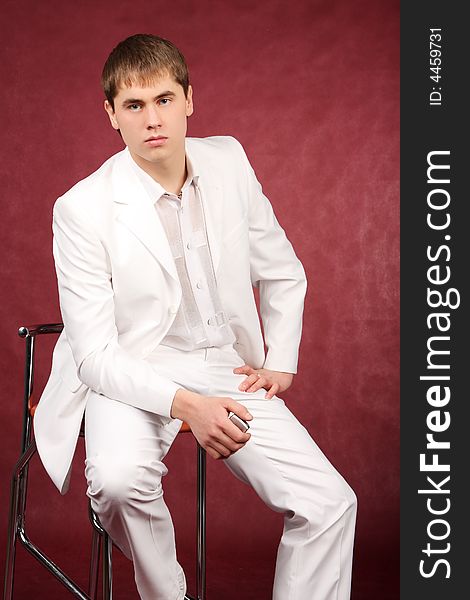 The portrait of the guy sitting on a chair in a white suit which holds a mobile phone tube in the right hand. The portrait of the guy sitting on a chair in a white suit which holds a mobile phone tube in the right hand