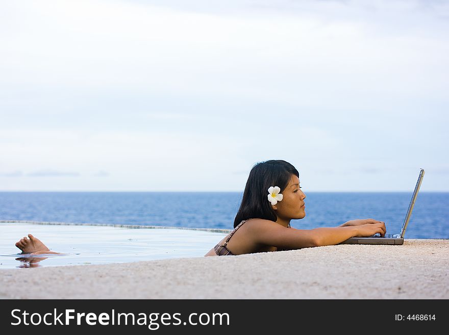 Attractive young Asian business woman working remotely on a laptop computer beside an infinity pool at a private tropical resort paradise vacation. Room for text. Attractive young Asian business woman working remotely on a laptop computer beside an infinity pool at a private tropical resort paradise vacation. Room for text.