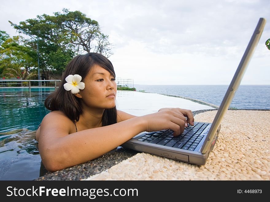 Attractive young Asian business woman working remotely on a laptop computer beside an infinity pool at a private tropical resort paradise vacation. Room for text. Attractive young Asian business woman working remotely on a laptop computer beside an infinity pool at a private tropical resort paradise vacation. Room for text.