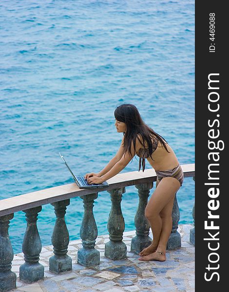 Attractive young Asian business woman working remotely on a laptop computer beside a balcony railing overlooking the ocean at a private tropical resort paradise vacation. Room for text. Attractive young Asian business woman working remotely on a laptop computer beside a balcony railing overlooking the ocean at a private tropical resort paradise vacation. Room for text.