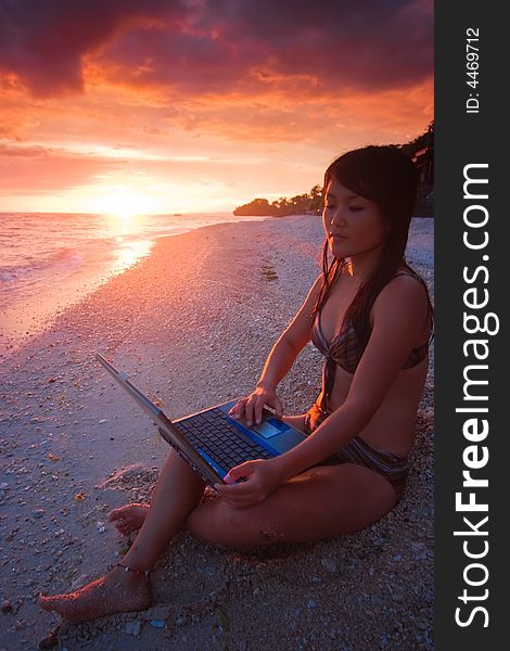 Attractive young Asian business woman working remotely on a laptop computer on a private white sand beach beside the ocean at tropical resort paradise vacation with beautiful red sunset background. Room for text. Attractive young Asian business woman working remotely on a laptop computer on a private white sand beach beside the ocean at tropical resort paradise vacation with beautiful red sunset background. Room for text.