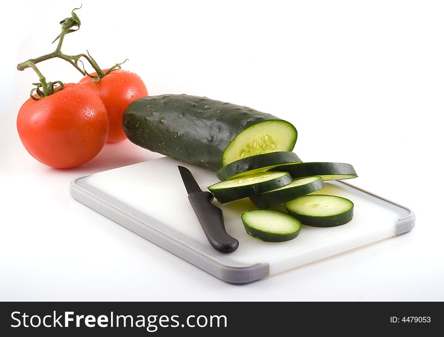 Slices of cucumber rest on a cutting board with two tomatoes sitting nearby. Slices of cucumber rest on a cutting board with two tomatoes sitting nearby.