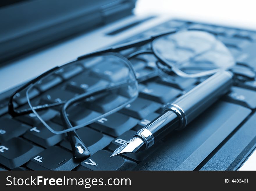 A pair of glasses and a fountain pen on a laptop on blue. Symbol of business working in office. Soft focus. A pair of glasses and a fountain pen on a laptop on blue. Symbol of business working in office. Soft focus.