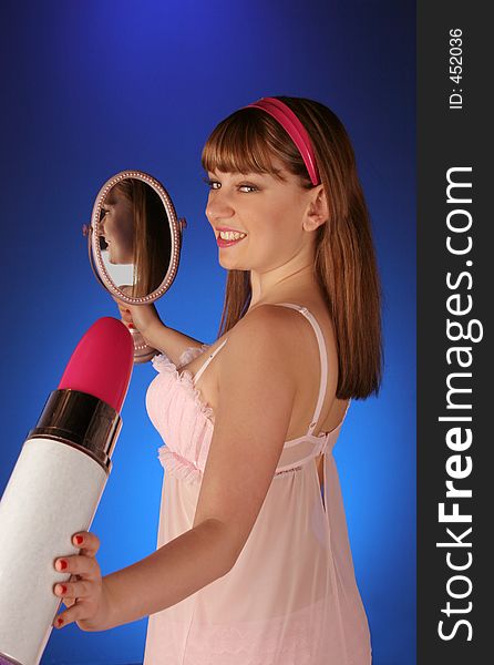 Teenager holding large lipstick and small hand mirror on blue backdrop looking in camera. Teenager holding large lipstick and small hand mirror on blue backdrop looking in camera
