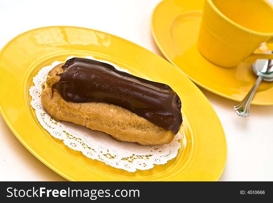 A chocolate eclair on yellow plate with coffee cup and saucer.