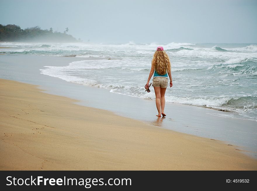 A young woman walks alone on the beach carrying her sandals and wearing a pink hat. A young woman walks alone on the beach carrying her sandals and wearing a pink hat