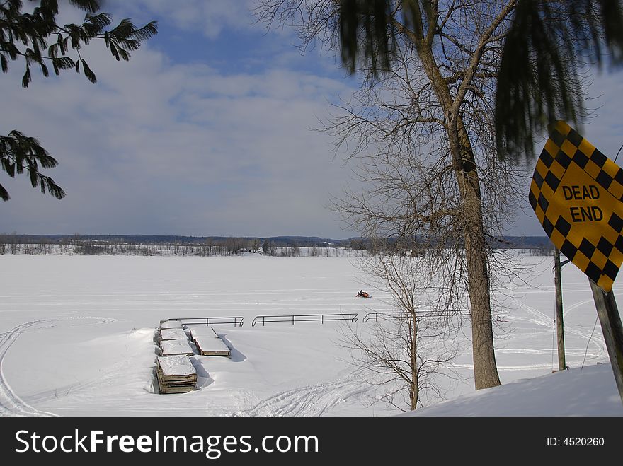 Dead end sign on a snowy lane.  A snowmobile passes by a boat dock on a frozen river.