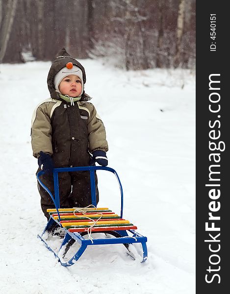 Baby in green coat standing near sled in snow-covered park. Baby in green coat standing near sled in snow-covered park