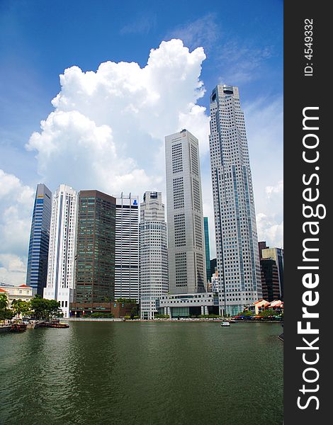 Towering skyscrapers loom over a reflective river and surrounded by huge clouds.