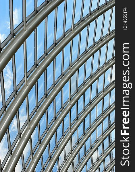 Silver metal curved roof joists in a conservatory with glass panes in between and a blue sky and clouds beyond. Silver metal curved roof joists in a conservatory with glass panes in between and a blue sky and clouds beyond.