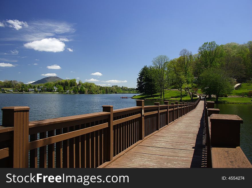 A footbridge leading across a small lake with trees and a blue sky in the background