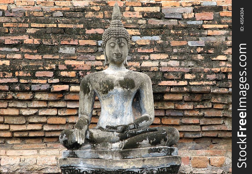 A seated concrete Buddha statue in front of an ancient brick wall in sukhothai National Park.  Sukhothai, Thailand.