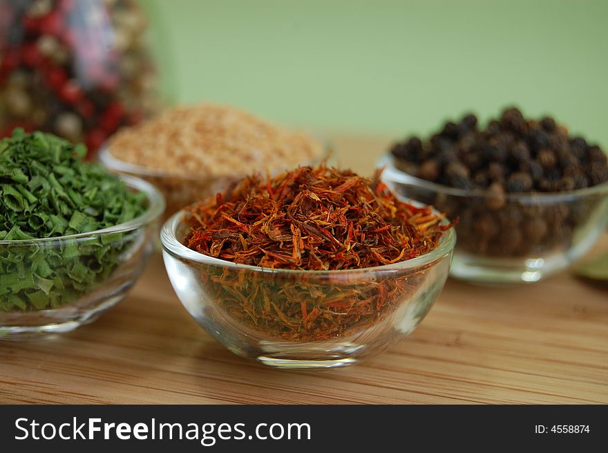 Glass containers with saffron, mustard seed, black pepper and chive and a bottle of mixed pepper on a wooden cutting desk with a green background. Glass containers with saffron, mustard seed, black pepper and chive and a bottle of mixed pepper on a wooden cutting desk with a green background