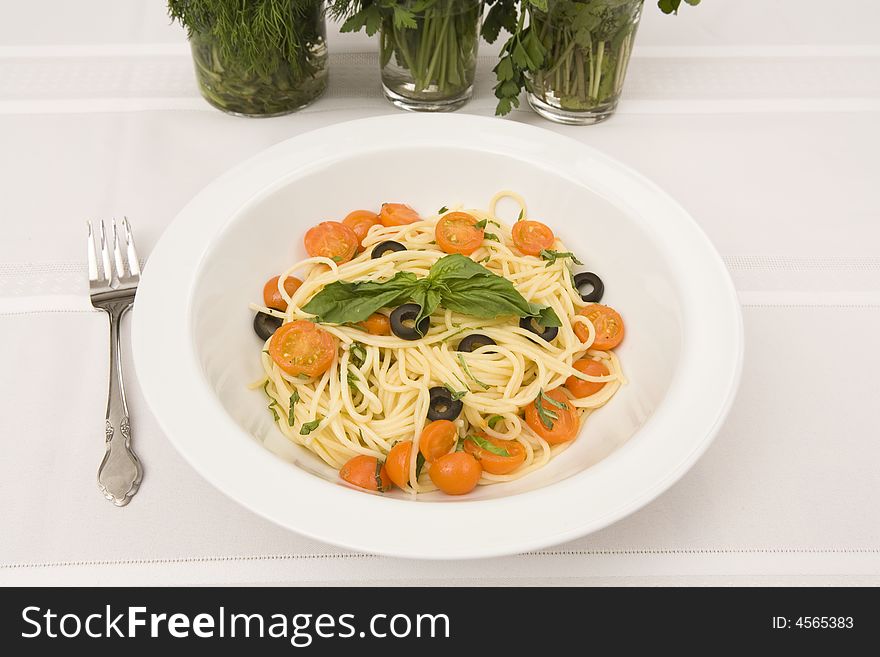 A table setting with a serving bowl of a spaghetti with tomatoes and black olives entree. The table cloth is white, the herbs in glasses are a decoration in the background.