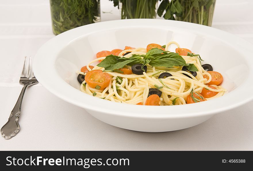 A table setting with a serving bowl of a spaghetti with tomatoes and black olives entree.  The table top is white and there are three decorative glasses with herbs in the background. A table setting with a serving bowl of a spaghetti with tomatoes and black olives entree.  The table top is white and there are three decorative glasses with herbs in the background.