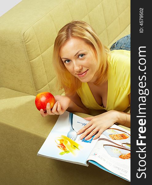 The girl lays on a sofa, reads the magazine and eats an apple. The girl lays on a sofa, reads the magazine and eats an apple