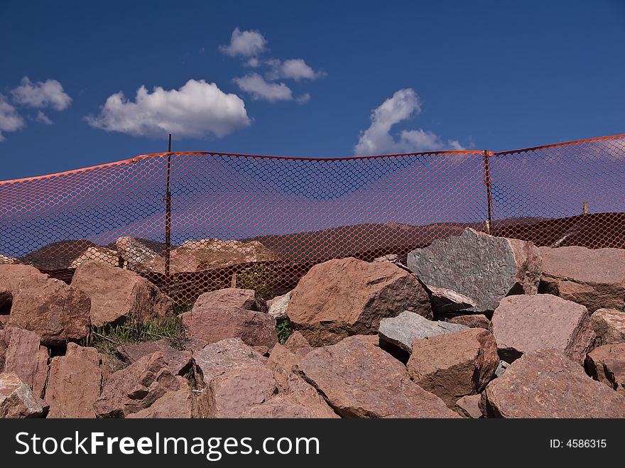 Orange construction fence above pile of large boulders with blue sky and white puffy clouds. Orange construction fence above pile of large boulders with blue sky and white puffy clouds