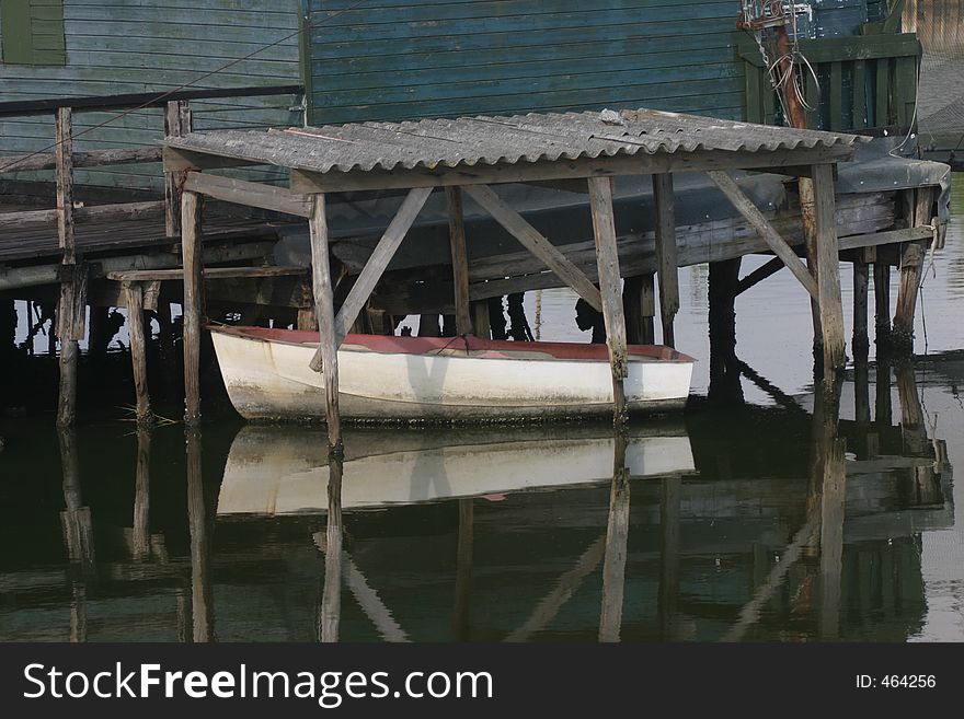 A small white boat under an old makeshift roof, with the scene reflecting in the calm water of the harbor. A small white boat under an old makeshift roof, with the scene reflecting in the calm water of the harbor