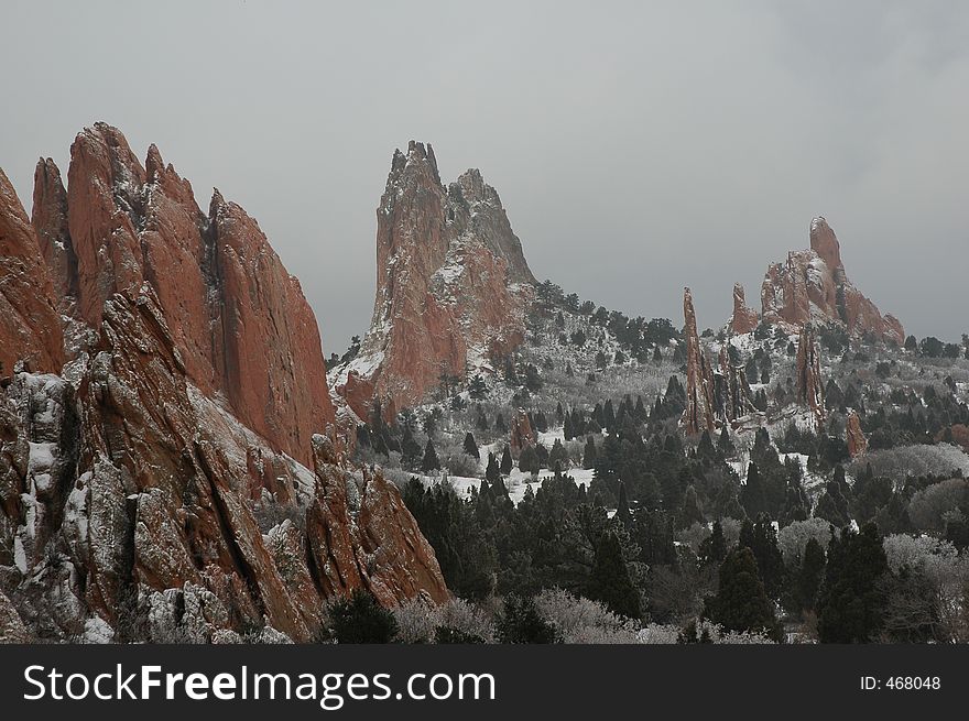 Image depicts a landscape scene in winter, with pines and spiked red rock formations in Colorado Spring's Garden of the Gods preserve. Image depicts a landscape scene in winter, with pines and spiked red rock formations in Colorado Spring's Garden of the Gods preserve