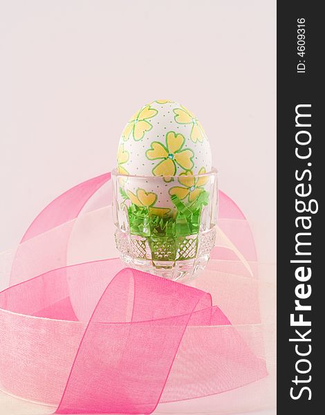 A single hand decorated Easter egg sits in a crystal cup surrounded by pink and yellow organza ribbon over a pale pink background. A single hand decorated Easter egg sits in a crystal cup surrounded by pink and yellow organza ribbon over a pale pink background.