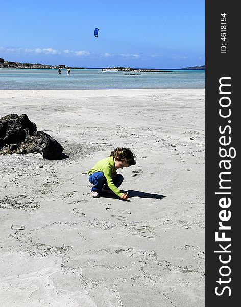 A child drawing on the sand of elafonisi beach in crete with the sea and surfing men as background