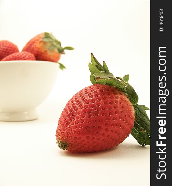 Image of a bright red strawberry in the foreground, and a small white bowl with other strawberries in the background.  White background.  Vertical orientation. Image of a bright red strawberry in the foreground, and a small white bowl with other strawberries in the background.  White background.  Vertical orientation.