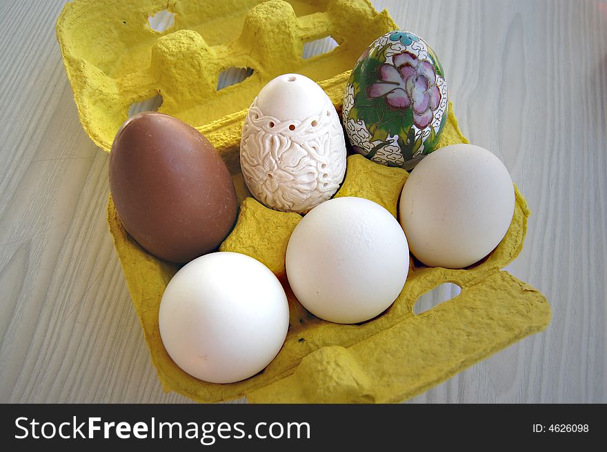 A chocolate egg, a chinese decorated porcelain egg, a hand carved meerschaum egg and three white eggs in a yellow egg box on white and gray oak laminate. A chocolate egg, a chinese decorated porcelain egg, a hand carved meerschaum egg and three white eggs in a yellow egg box on white and gray oak laminate