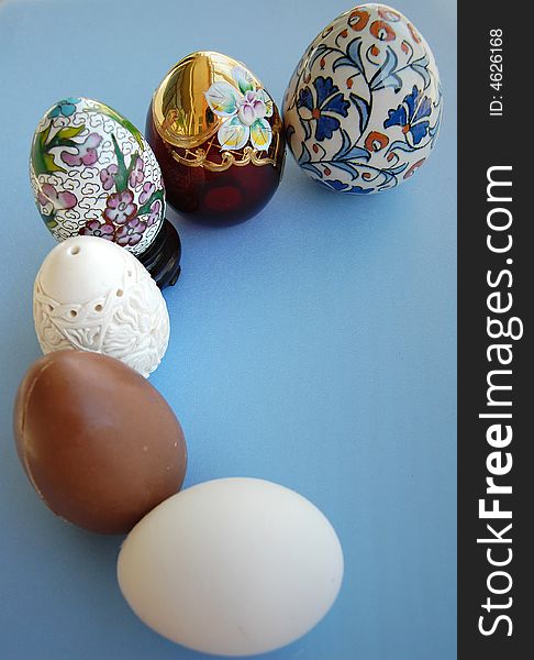 A chocolate egg, a Chinese decorated porcelain egg, a hand carved meerschaum egg, a burgundy and gold glass egg and a white eggs on blue background together. A chocolate egg, a Chinese decorated porcelain egg, a hand carved meerschaum egg, a burgundy and gold glass egg and a white eggs on blue background together