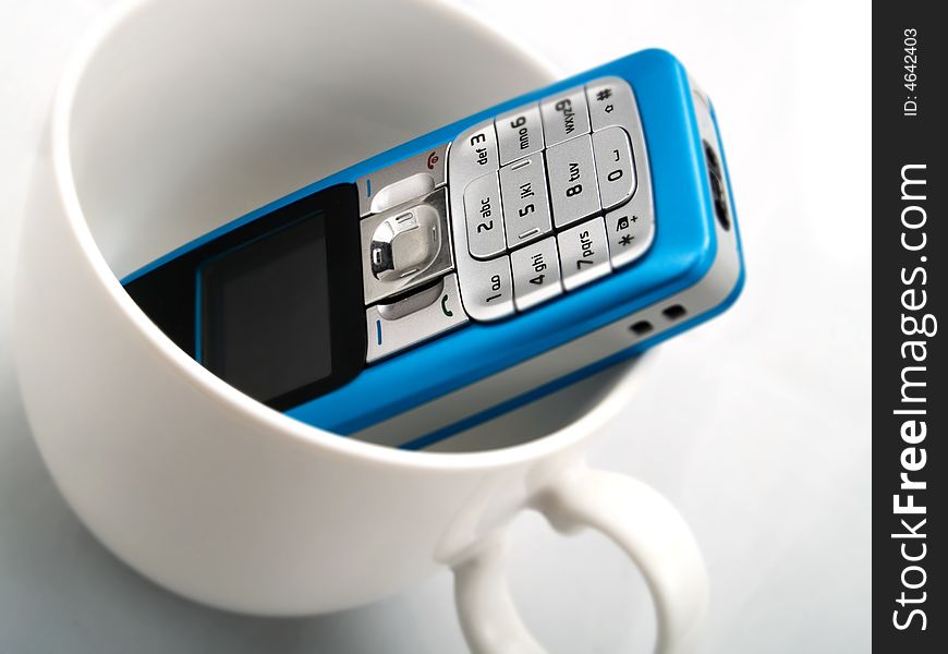 A light blue mobile phone staying in a white cup. A light blue mobile phone staying in a white cup