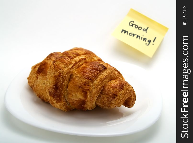 A fresh croissant in a white plate and a yellow note with this text: 'Good morning!'. A fresh croissant in a white plate and a yellow note with this text: 'Good morning!'