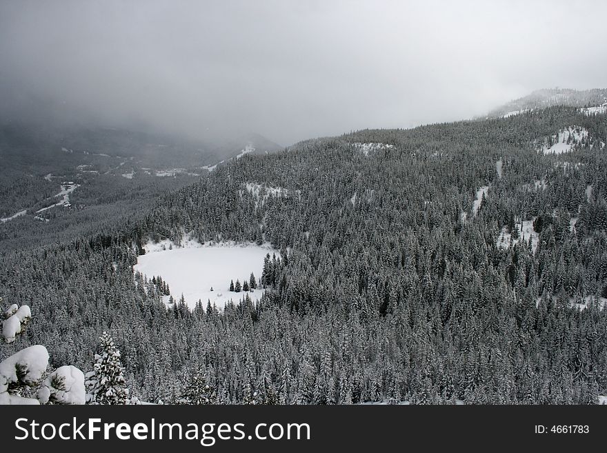 A frozen lake surrounded by snow covered pines is nestled in an all white snowy winter wonderland. A frozen lake surrounded by snow covered pines is nestled in an all white snowy winter wonderland