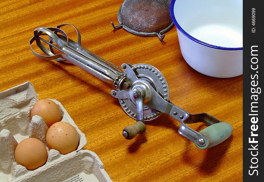An old and well-used manual egg-beater lying on a table, with related items. An old and well-used manual egg-beater lying on a table, with related items.