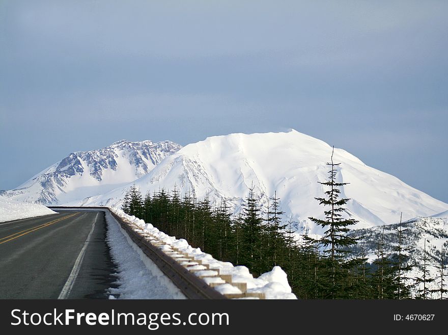 Paved road with snow packed guardrail, the peak of snow covered mountain, blue sky, evergreen trees in foreground. Paved road with snow packed guardrail, the peak of snow covered mountain, blue sky, evergreen trees in foreground