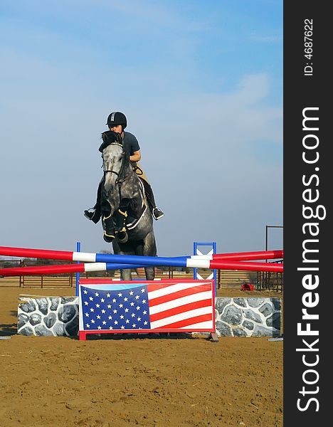 Young woman riding dappled grey horse over jump with painted Amercian flag. Young woman riding dappled grey horse over jump with painted Amercian flag.