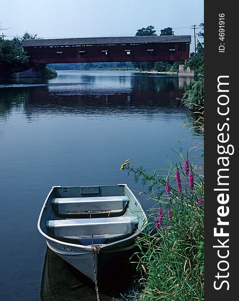 A row boat sits tied to the shore with a covered bridge in the background