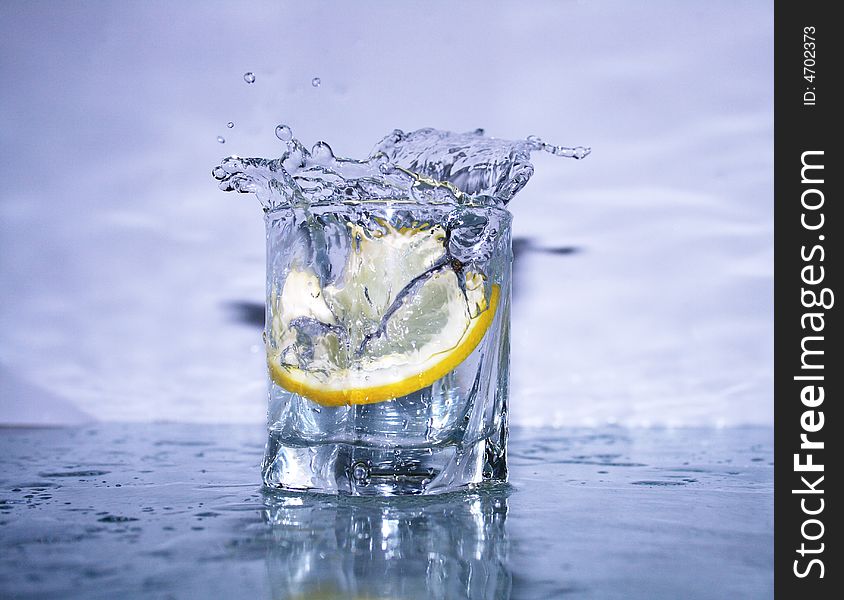 Glass of cold clear splashing water with falling lemon's slice on light-blue background. Glass of cold clear splashing water with falling lemon's slice on light-blue background