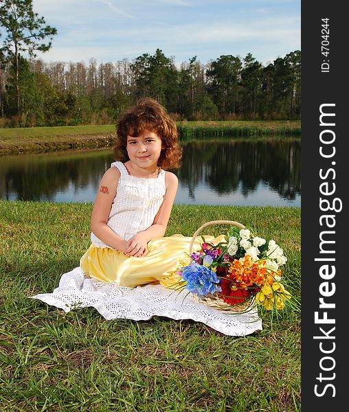 A pretty little  girl is sitting on the grass with the basket of flowers next to her on the lake background. A pretty little  girl is sitting on the grass with the basket of flowers next to her on the lake background