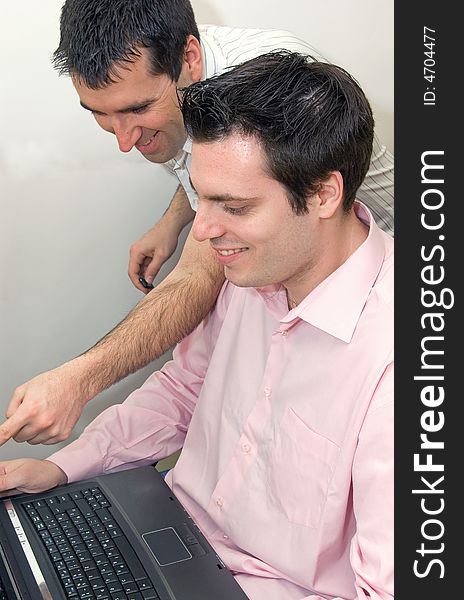 Two young men, working on a laptop computer, one sitting in his chair, and the other, standing next to him, both smiling. Two young men, working on a laptop computer, one sitting in his chair, and the other, standing next to him, both smiling