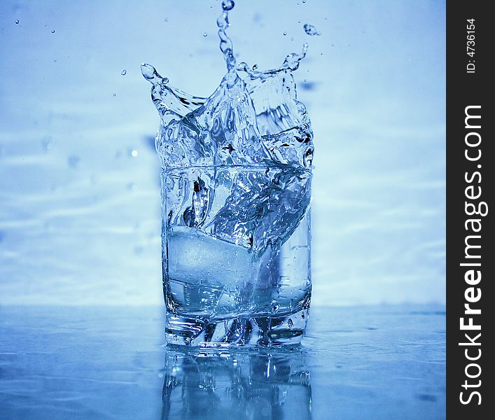 Glass of splashing water with ice on blue background