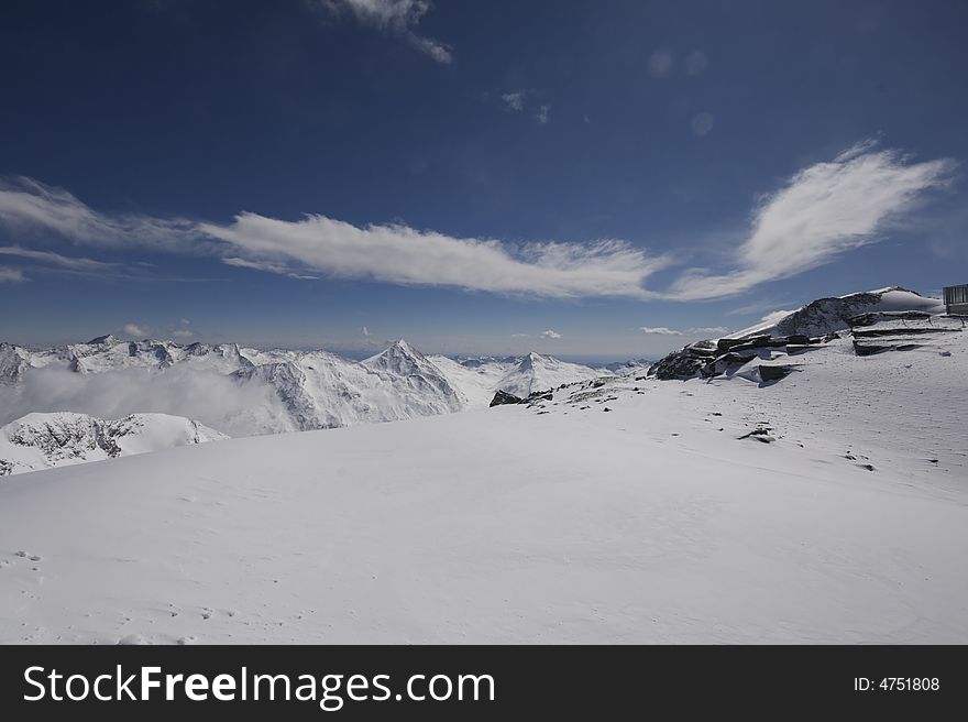 This photograph shows the top of a mountain in the resort of Saas-Fee in Switzerland. There are ski marks in the snow, and you can see down the valley over Saas-Fee and other mountain villages. This photograph shows the top of a mountain in the resort of Saas-Fee in Switzerland. There are ski marks in the snow, and you can see down the valley over Saas-Fee and other mountain villages.