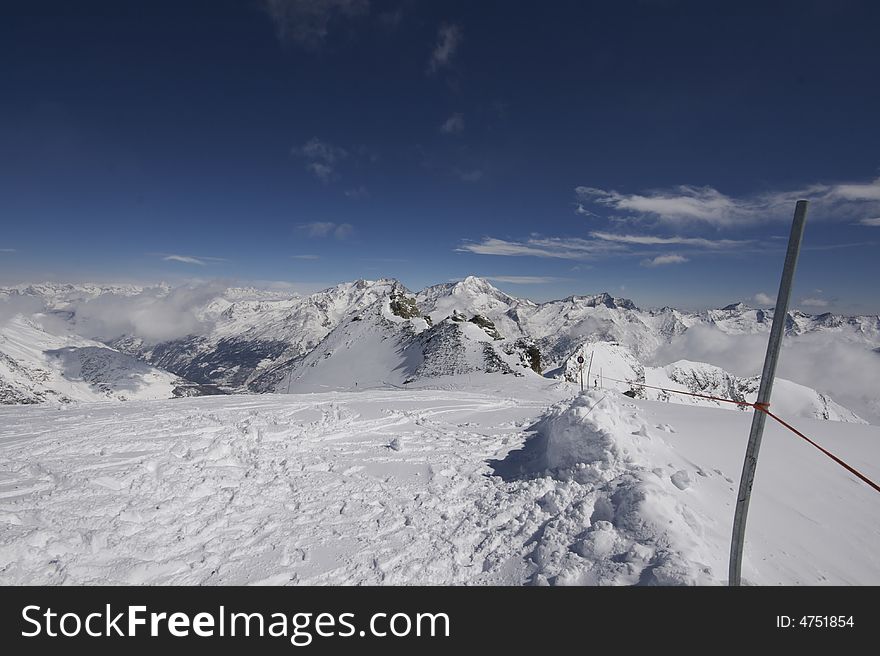 This photograph shows the top of a mountain in the resort of Saas-Fee in Switzerland. There are ski marks in the snow, and you can see down the valley over Saas-Fee and other mountain villages. This photograph shows the top of a mountain in the resort of Saas-Fee in Switzerland. There are ski marks in the snow, and you can see down the valley over Saas-Fee and other mountain villages.