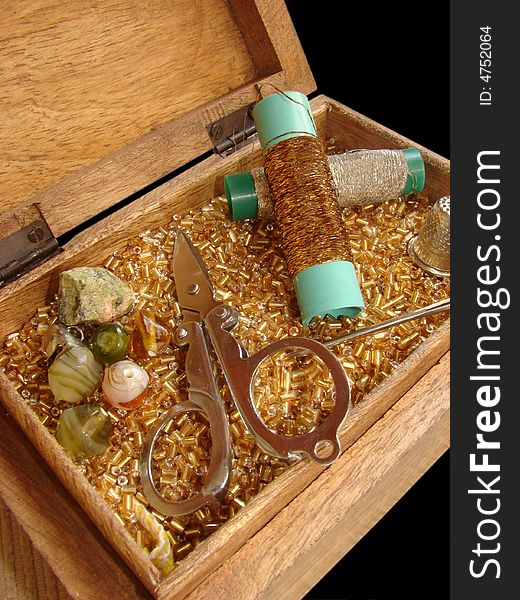 Sewing basket in golden colors, containing scissors, beads, stones, golden and silver thread, thimble and needle on a black background.