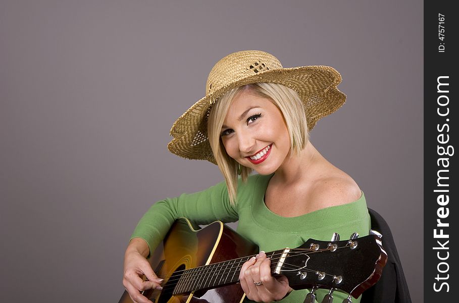 A blonde in a green blouse and straw hat playing the guitar and smiling. A blonde in a green blouse and straw hat playing the guitar and smiling