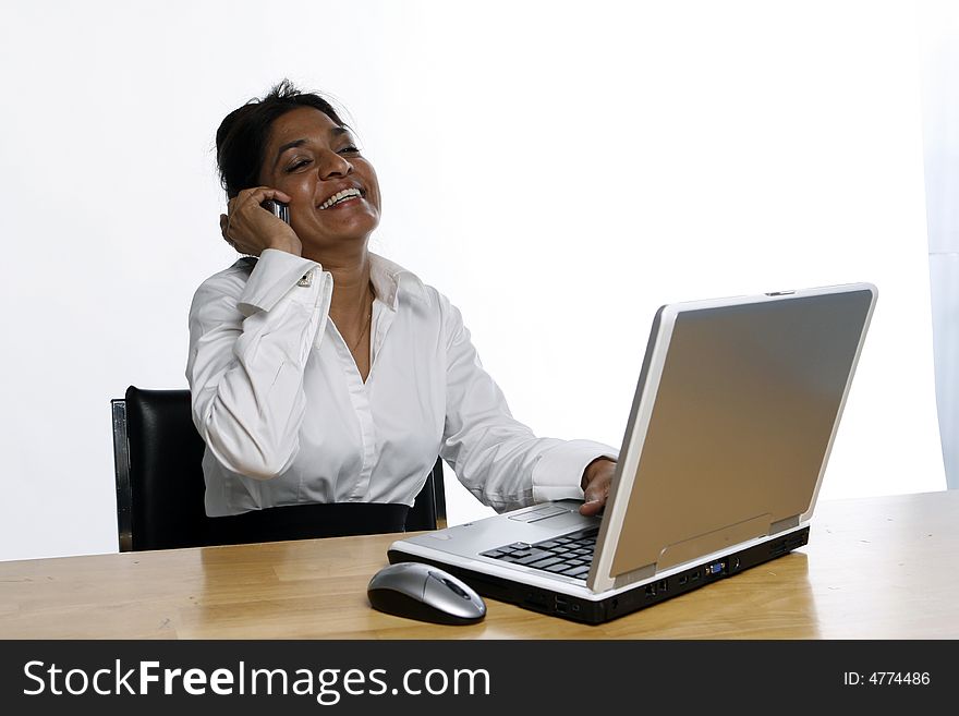 Indian business woman leaning back and laughing while on her phone sitting in front of her laptop. Indian business woman leaning back and laughing while on her phone sitting in front of her laptop