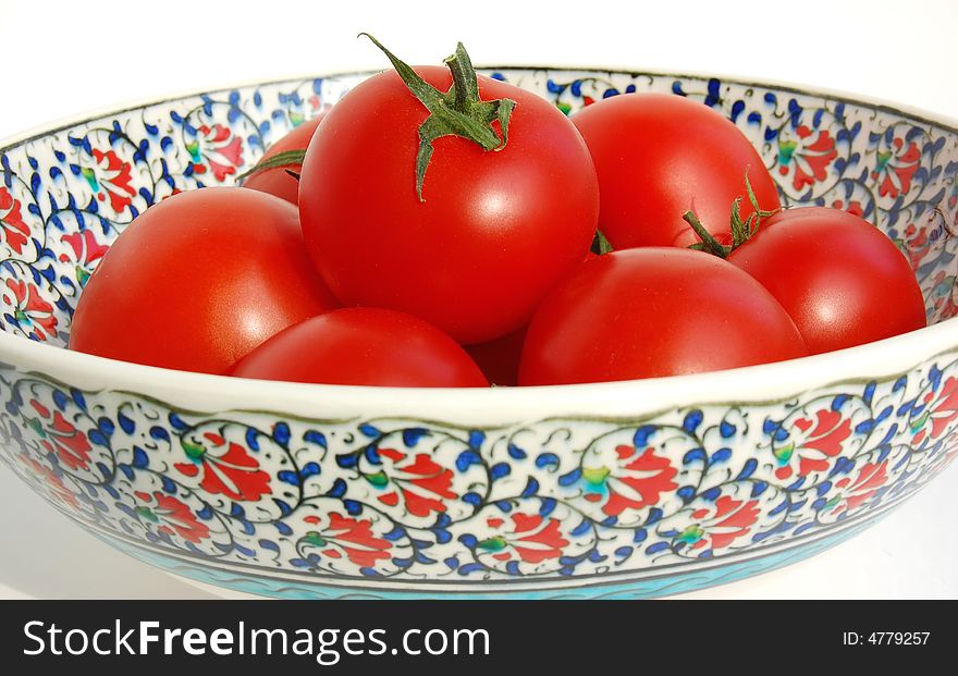 Glossy red ripe round small tomatos with stalks in a hand painted Iznik ceramic bowl with classical red, green, blue, turquoise carnation patterns on white. Glossy red ripe round small tomatos with stalks in a hand painted Iznik ceramic bowl with classical red, green, blue, turquoise carnation patterns on white.