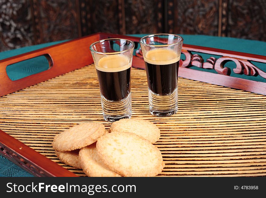 Freshly brewed espresso shots in shot glasses, on a serving plate with cookies. Shot from the side.