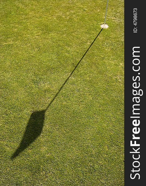 On the Putting green of a golf course the flag and pin or stick casting a shadow on the green. On the Putting green of a golf course the flag and pin or stick casting a shadow on the green