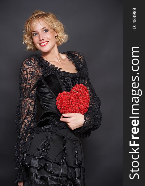The beautiful blonde in an evening dress holds in hands heart made of roses. Background is black. The beautiful blonde in an evening dress holds in hands heart made of roses. Background is black