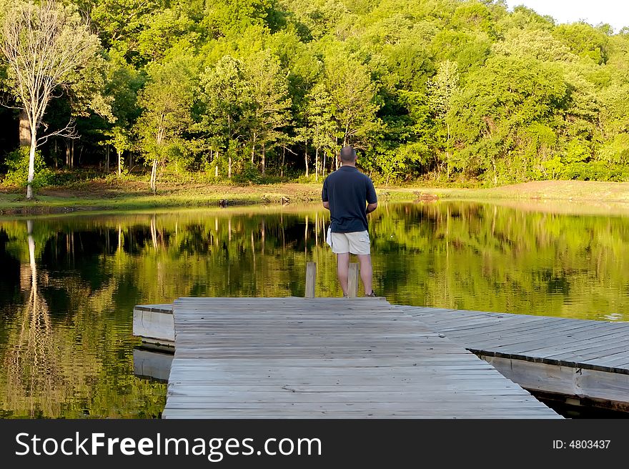 An image of a man fishing very early in the morning off a pier on a small pond